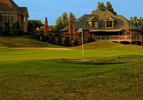 this color image is of a Putting Green on Professional Golf Course near Luxury Homes. the putting green has manicured grass or lawn with two sand traps around the outside of the putting green and putting hole. the grass is green and their are expensive houses or homes in the background. the photo was taken during the spring or summer. and the lighting is natural sunlight during the evening part of the day. there are also trees with leaves in the background.